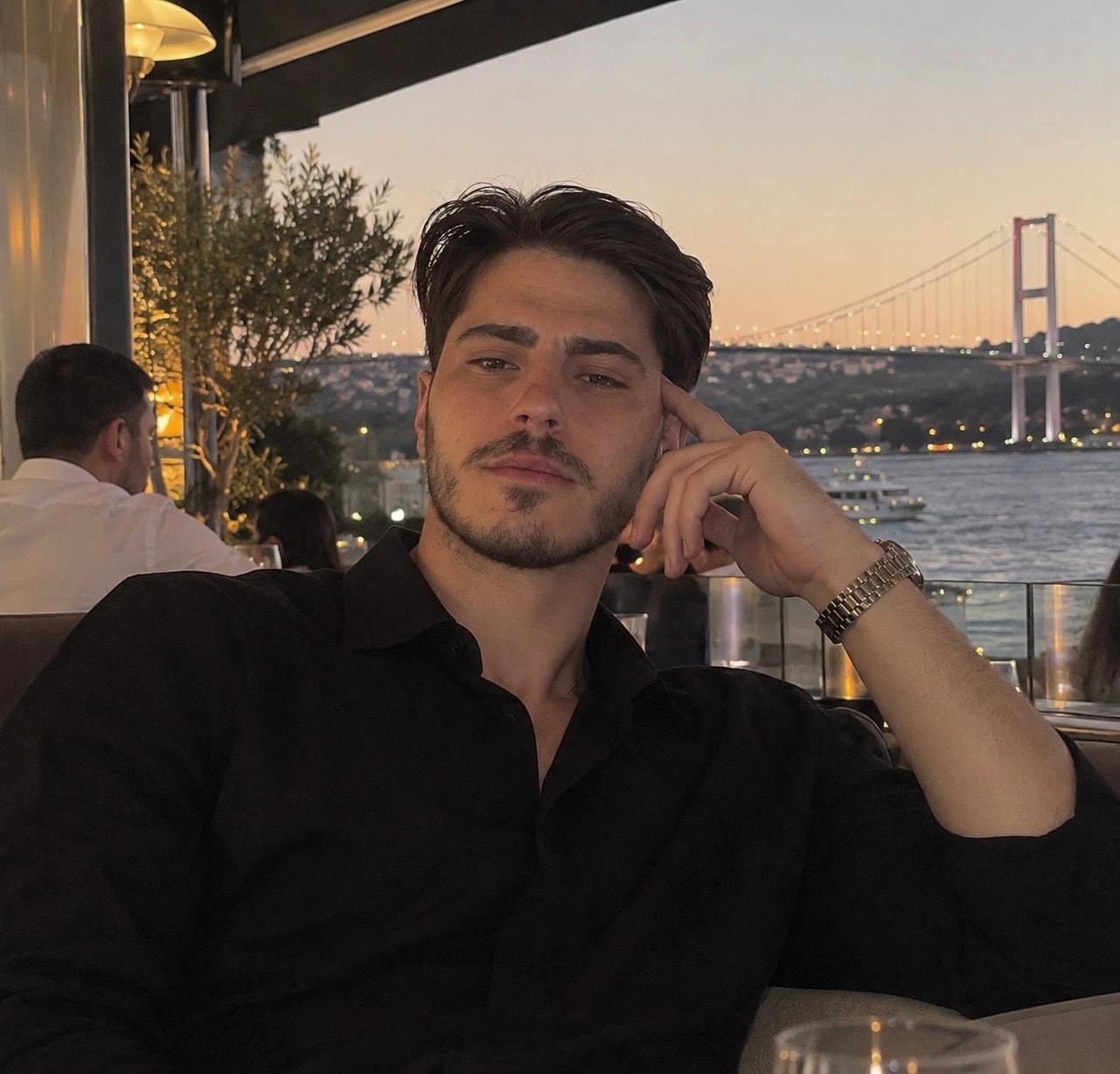 Mustafa; siyah gömlekle bir terasta oturuyor, arkasında gün batımında Boğaz Köprüsü.